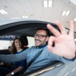 Um homem sorridente ao volante de um carro alugado, fazendo o sinal de "OK" com a mão, enquanto uma mulher sentada no banco do passageiro sorri.