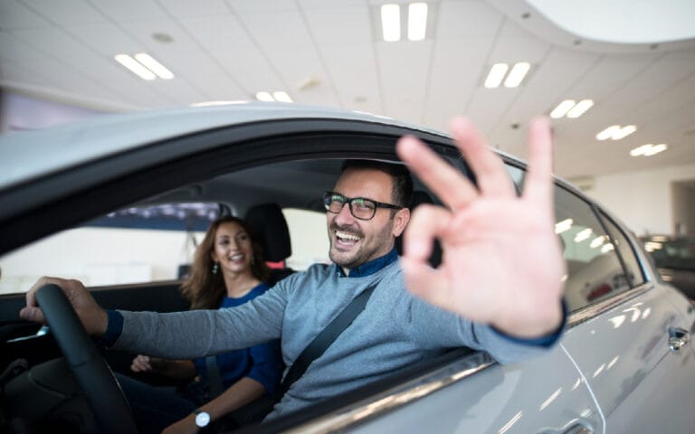 Um homem sorridente ao volante de um carro alugado, fazendo o sinal de "OK" com a mão, enquanto uma mulher sentada no banco do passageiro sorri.